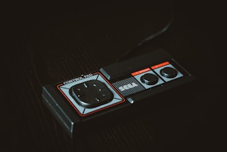 A vintage gaming controller featuring a rectangular design with a directional pad and two round buttons, labeled as 'SEGA' and 'Control Pad'. The controller is set against a dark background, highlighting its retro aesthetic.
