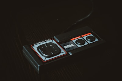 A vintage gaming controller featuring a rectangular design with a directional pad and two round buttons, labeled as 'SEGA' and 'Control Pad'. The controller is set against a dark background, highlighting its retro aesthetic.