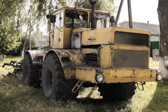 A large, yellow, rustic tractor sits on grassy terrain near a wooden house with a green roof. The machine appears weathered, with visible wear and rust. Its oversized tires and metal structure enhance its robust appearance. Nearby, trees cast dappled shadows on the vehicle, contributing to the rural atmosphere.