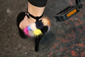 A person's foot wearing a high-heeled shoe with colorful pom-poms attached to the strap. The pom-poms are in shades of blue, pink, yellow, black, and orange. The background is a textured surface with hints of red and gray.
