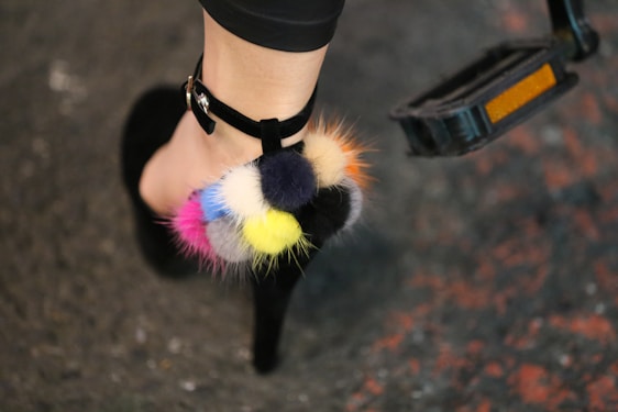 A person's foot wearing a high-heeled shoe with colorful pom-poms attached to the strap. The pom-poms are in shades of blue, pink, yellow, black, and orange. The background is a textured surface with hints of red and gray.