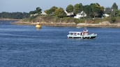 A small boat carrying passengers travels across a body of blue water. In the background, a lush green shoreline is dotted with white houses surrounded by numerous trees. A yellow and black striped buoy floats in the water, indicating navigation guidance.