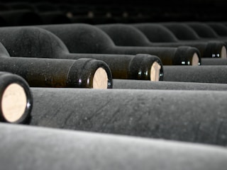 Several rows of dusty wine bottles are stored horizontally, their corks facing outwards. The bottles are arranged neatly in a dimly lit environment, suggesting a wine cellar or storage area.