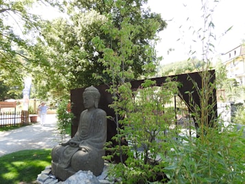 A serene garden scene featuring a large stone Buddha statue surrounded by lush greenery and trees, with a wooden structure partially visible behind it. Soft light filters through the leaves, creating a peaceful atmosphere. In the background, a walkway and a person walking can be seen.
