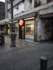 An urban street scene featuring a storefront with a bright red signage that reads 'Iwo's' on a partially overcast day. The building facade shows wear, with multiple air conditioning units attached. A motor scooter is parked on the left side, and several posters are stuck to a lamppost in the foreground. Checkerboard patterns adorn parts of the shop's exterior, and interior lights are on.