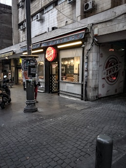 An urban street scene featuring a storefront with a bright red signage that reads 'Iwo's' on a partially overcast day. The building facade shows wear, with multiple air conditioning units attached. A motor scooter is parked on the left side, and several posters are stuck to a lamppost in the foreground. Checkerboard patterns adorn parts of the shop's exterior, and interior lights are on.