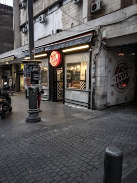 An urban street scene featuring a storefront with a bright red signage that reads 'Iwo's' on a partially overcast day. The building facade shows wear, with multiple air conditioning units attached. A motor scooter is parked on the left side, and several posters are stuck to a lamppost in the foreground. Checkerboard patterns adorn parts of the shop's exterior, and interior lights are on.