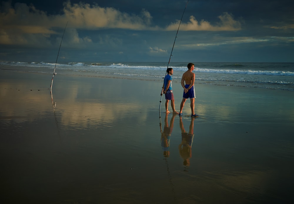 two boy standing beside fishing rod stump on shoreline at daytime