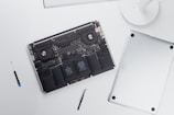 An open laptop with its back cover removed reveals internal components such as the battery, fans, and circuitry. Next to the laptop, there is a screwdriver and a tool, possibly used for disassembly.