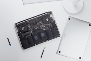 An open laptop with its back cover removed reveals internal components such as the battery, fans, and circuitry. Next to the laptop, there is a screwdriver and a tool, possibly used for disassembly.