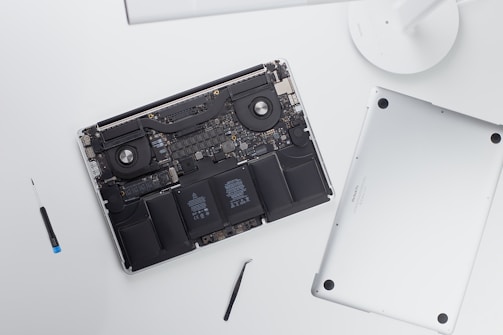 An open laptop with its back cover removed reveals internal components such as the battery, fans, and circuitry. Next to the laptop, there is a screwdriver and a tool, possibly used for disassembly.