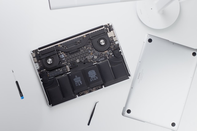 An open laptop with its back cover removed reveals internal components such as the battery, fans, and circuitry. Next to the laptop, there is a screwdriver and a tool, possibly used for disassembly.