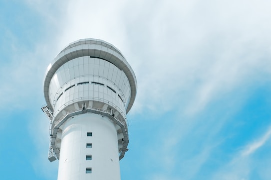 A modern, tall white control tower with a cylindrical shape stands against a bright blue sky with wispy clouds. The structure features multiple observation windows near the top.