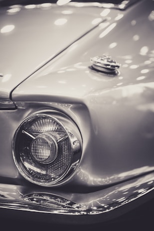 A close-up of a vintage car headlight with a chrome finish and sleek, curved design. The image features a gas cap on the hood, with multiple reflections from lights, creating a polished and reflective surface.