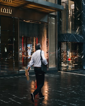 A person wearing a white shirt and dark jeans walks on a wet pavement in front of a luxury storefront. The reflection of lights on the ground suggests it has recently rained. The store displays luxurious items with an elegant interior design and the person is holding a rolled-up poster or paper in one hand while carrying a black shoulder bag.