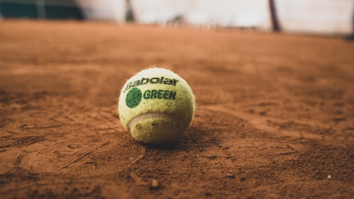 A tennis ball with visible branding lies on a reddish clay court, as shown by the textured surface beneath it. The focus is primarily on the ball, with the background softly blurred.