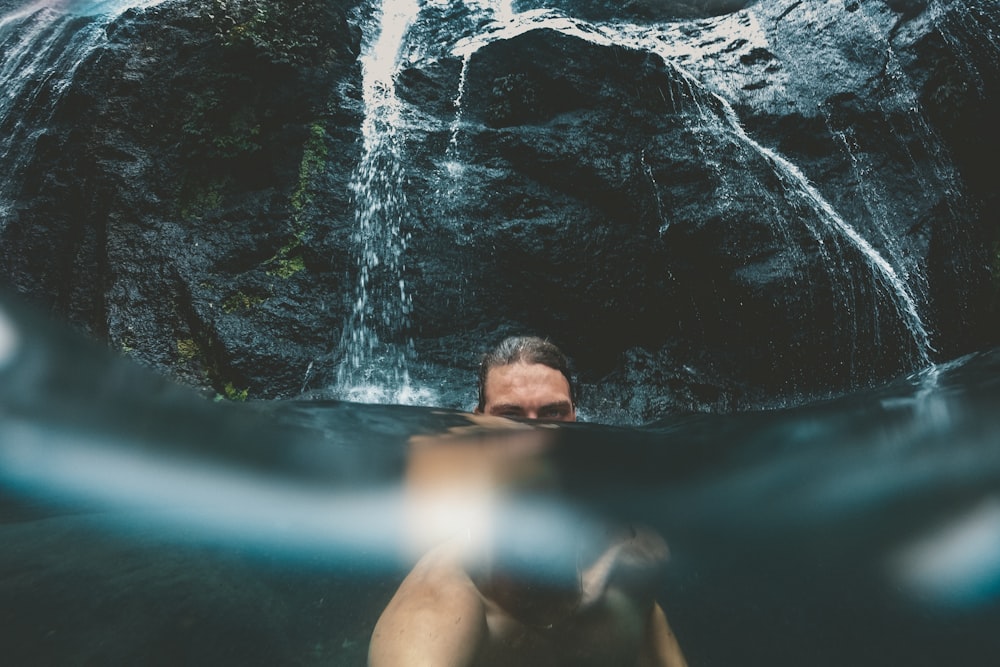 man talking a selfie half underwater in front of waterfalls
