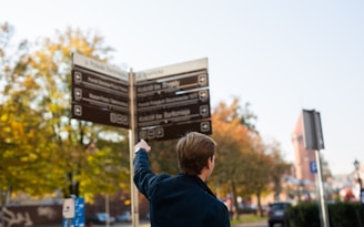 A person wearing a dark jacket is pointing at a directional signpost. The signpost displays multiple signs with directions and information in various languages. The background features trees with autumn leaves, hinting at a park or urban setting. The sky is clear, and the atmospheric lighting suggests a sunny day.