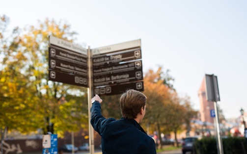 A person wearing a dark jacket is pointing at a directional signpost. The signpost displays multiple signs with directions and information in various languages. The background features trees with autumn leaves, hinting at a park or urban setting. The sky is clear, and the atmospheric lighting suggests a sunny day.