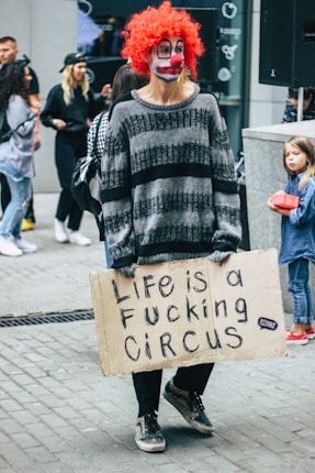 A person dressed as a clown with a red wig and painted face stands on a city street holding a cardboard sign with bold black letters saying 'Life is a Fucking Circus.' Passersby, including a child holding a red object, are visible in the background.