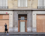 A person in a dark coat is walking past a boarded-up building with the sign 'AGENCE O' above the entrance. The windows are covered with plywood, and there is red graffiti on one of the panels. The scene appears to be urban, and the street is littered with leaves.