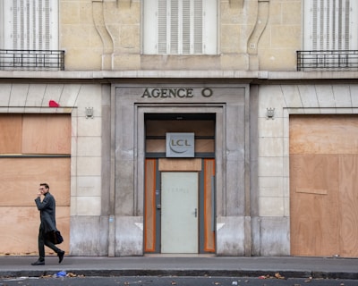 A person in a dark coat is walking past a boarded-up building with the sign 'AGENCE O' above the entrance. The windows are covered with plywood, and there is red graffiti on one of the panels. The scene appears to be urban, and the street is littered with leaves.