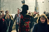 A person wearing traditional Scottish attire, including a red tartan kilt and a black feathered cap, stands in the middle of a crowded street. This individual is playing bagpipes, surrounded by people walking in various directions.