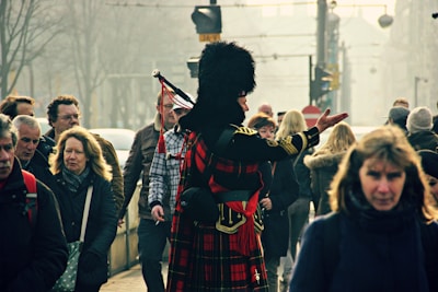 A person wearing traditional Scottish attire, including a red tartan kilt and a black feathered cap, stands in the middle of a crowded street. This individual is playing bagpipes, surrounded by people walking in various directions.