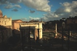 Ancient Roman ruins are under a partly cloudy sky, bathed in warm sunlight. Architectural structures like columns and a triumphal arch are prominent, with trees and hills visible in the background. The scene suggests a combination of decay and historical grandeur.