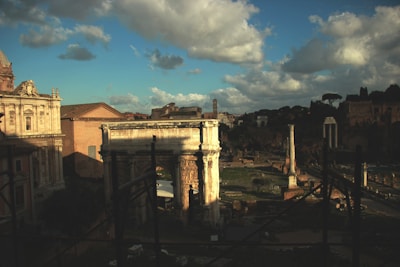 Ancient Roman ruins are under a partly cloudy sky, bathed in warm sunlight. Architectural structures like columns and a triumphal arch are prominent, with trees and hills visible in the background. The scene suggests a combination of decay and historical grandeur.