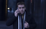 A person wearing a dark coat and scarf is holding a phone to their ear, set against a dimly lit urban background. Their expression appears focused and slightly concerned.