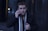 A person wearing a dark coat and scarf is holding a phone to their ear, set against a dimly lit urban background. Their expression appears focused and slightly concerned.