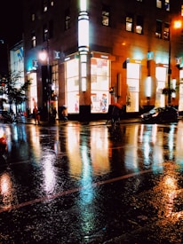 A nighttime cityscape featuring a brightly lit corner building with large windows, reflecting its lights on the wet pavement below. Pedestrians with umbrellas walk across the street, and several cars are parked nearby. Streetlights add to the illumination, creating colorful reflections on the rainy surface.