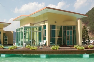A modern house with large glass windows and doors surrounded by a wooden deck. The building is situated over a body of water, giving it a floating appearance. There are potted plants on the deck and a chair for relaxing. The backdrop includes a forested hillside under a blue sky with some clouds.