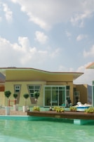 A modern house with a geometric design stands near a swimming pool. There are several potted trees and plants arranged in front of the house, enhancing the aesthetic appeal. The sky is clear with a few clouds, and the sun casts a bright light over the scene.