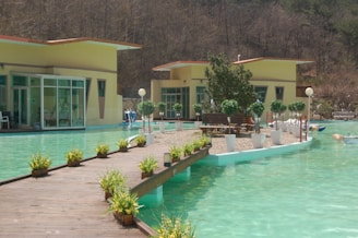 A serene outdoor setting featuring a light turquoise water pool with a wooden walkway lined with small potted plants leading to a small island. The island has a seating area surrounded by white plant pots with green shrubs. In the background, modern flat-roofed buildings with large windows are visible, set against a backdrop of hilly terrain.
