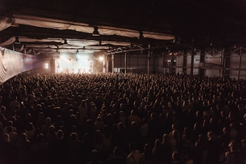 A large crowd of people gathered in a dimly lit indoor venue facing a brightly lit stage. The venue appears to have a high ceiling with industrial lighting fixtures and the stage is set up with various lights and equipment, producing a bright, focused light towards the audience.