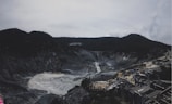 A volcanic crater with rocky and barren surroundings is visible. Sulfurous fumes emanate from the center, creating a distinctive color and atmosphere. In the foreground, structures and pathways accommodate visitors, while mountains form the background under a cloudy sky.