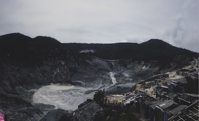A volcanic crater with rocky and barren surroundings is visible. Sulfurous fumes emanate from the center, creating a distinctive color and atmosphere. In the foreground, structures and pathways accommodate visitors, while mountains form the background under a cloudy sky.