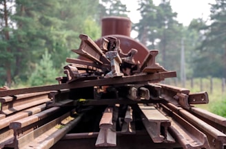 A stack of rusty metal railway tracks is arranged on a flatbed, set against a backdrop of lush green trees. The tracks are piled atop one another, showcasing signs of age and weathering.
