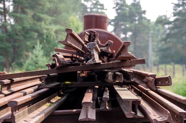 A stack of rusty metal railway tracks is arranged on a flatbed, set against a backdrop of lush green trees. The tracks are piled atop one another, showcasing signs of age and weathering.