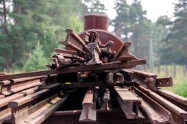 A stack of rusty metal railway tracks is arranged on a flatbed, set against a backdrop of lush green trees. The tracks are piled atop one another, showcasing signs of age and weathering.
