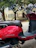 A red scooter is parked on the side of the street. It has a sleek design with chrome details on the handlebars and mirrors. The brown leather seat appears comfortable and stylish. In the background, there is an older stone building with arched windows and trees providing shade.