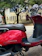 A red scooter is parked on the side of the street. It has a sleek design with chrome details on the handlebars and mirrors. The brown leather seat appears comfortable and stylish. In the background, there is an older stone building with arched windows and trees providing shade.