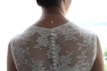 A woman is wearing a lace dress with a floral pattern. The back of the dress features a row of white buttons running down the spine. The dress material is sheer, allowing light to pass through, creating a delicate and elegant appearance. A silver necklace with a small pendant is visible on her neck. Her hair is partially visible, pulled back neatly.