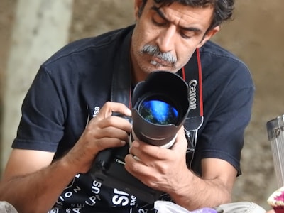 A person is closely examining a Canon DSLR camera with attention. The camera lens reflects what appears to be a distant scene or light source, giving it a captivating glow.