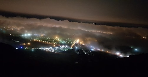 Thick clouds hover over a landscape at night, with a series of visible lights from urban areas below. These lights form a network of illuminated paths and clusters, suggesting streets and buildings. The scene is partially obscured by fog, creating a mysterious and ethereal atmosphere.
