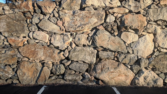 A wall constructed from large, irregularly shaped stones stacked together. The stones vary in size and color, primarily featuring shades of gray and brown. There is a parking lot at the base with visible white lines on the asphalt.