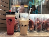 Two beverages in plastic cups with lids and straws are placed on a surface. The left cup contains a pink-red drink, possibly a fruit juice, with a black lid and a heart on a stick. The right cup holds a creamy beige-colored drink with ice cubes and a transparent straw. In the background, there are additional cups filled with strawberries and packaging materials.