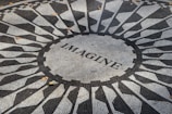 A circular mosaic pattern featuring geometric shapes in black and white, with the word 'IMAGINE' written at the center. The mosaic is situated outdoors and a few fallen leaves can be seen scattered across the surface.
